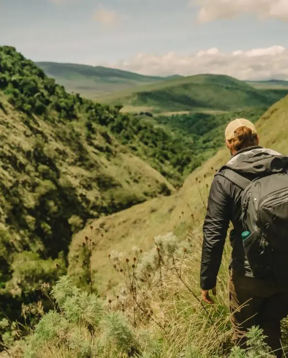 Two people hiking along a narrow grassy trail through a steep green valley, carrying backpacks and overlooking rolling hills and layered highland landscapes under a bright sky.