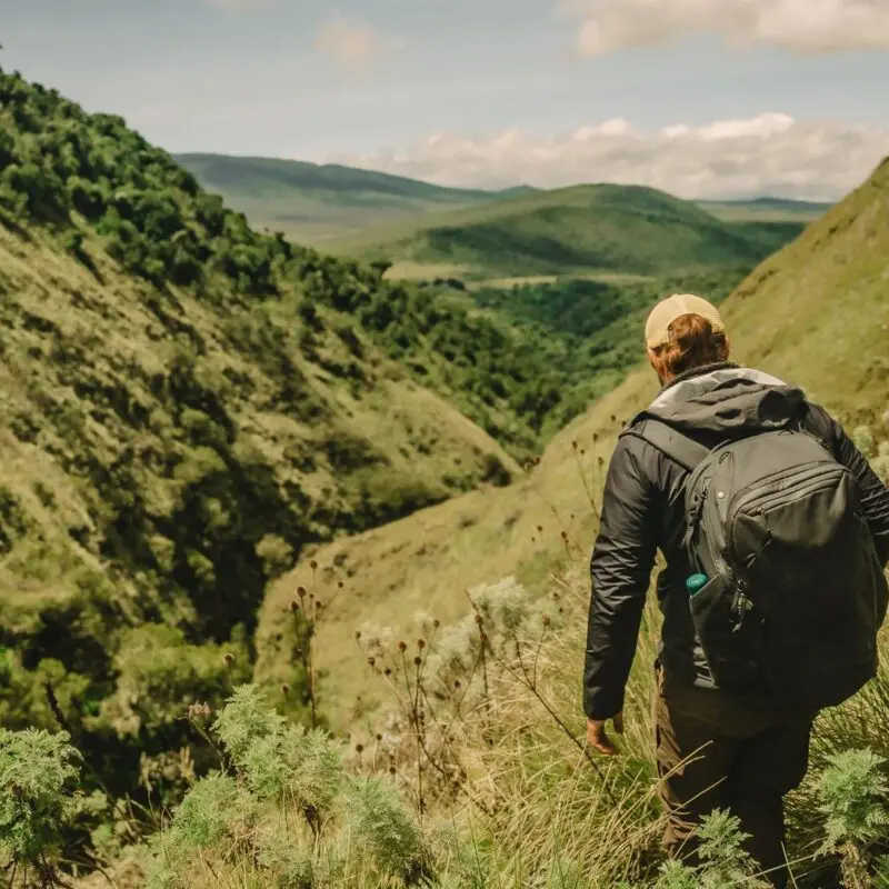 Two people hiking along a narrow grassy trail through a steep green valley, carrying backpacks and overlooking rolling hills and layered highland landscapes under a bright sky.