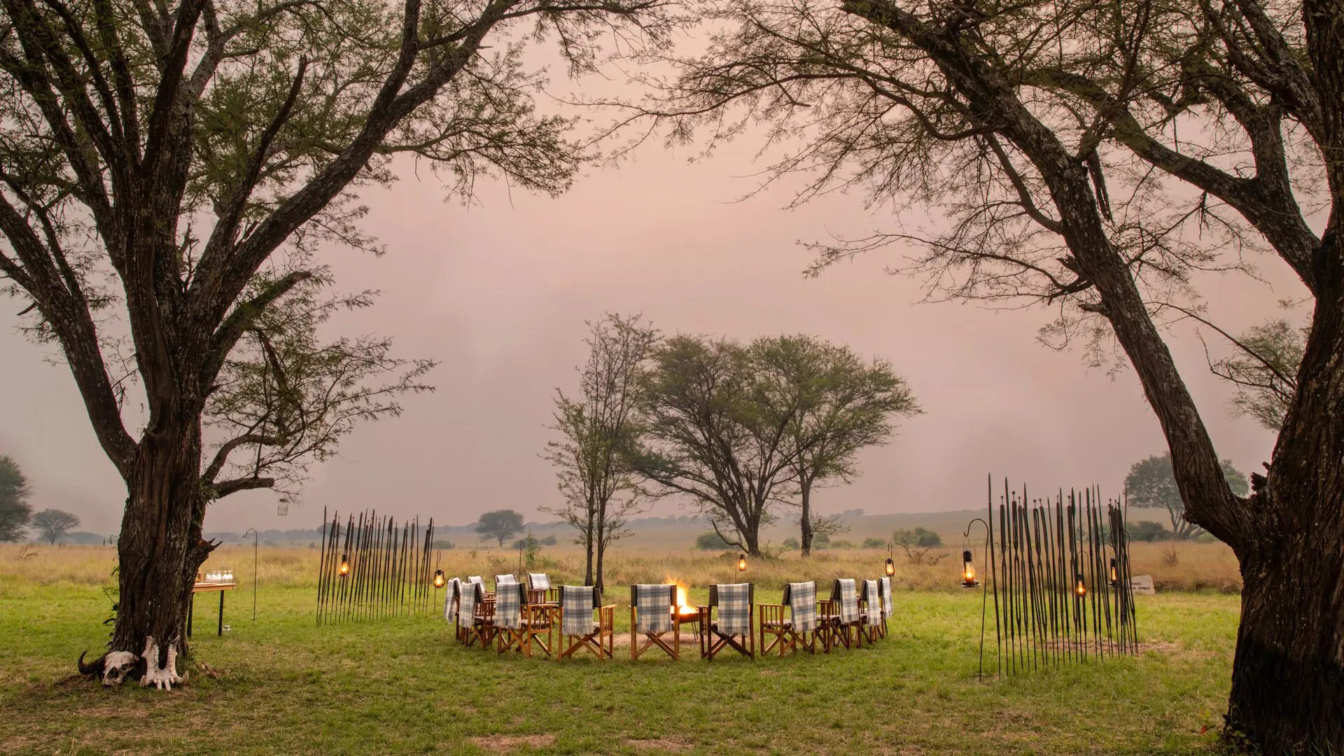 A circular arrangement of safari chairs surrounds a glowing campfire on open grassland, framed by tall acacia trees and lanterns, with the savannah stretching into the distance at dusk.