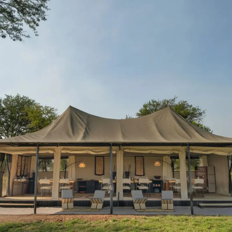 Ubuntu Migration Camp's spacious open-sided safari dining tent with neatly arranged tables and chairs, canvas walls, and modern furnishings, set among trees and grassland under a clear sky.