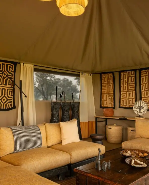 Interior of Ubuntu Migration Camp's lounge featuring cushioned sofas, woven textiles and wall art, soft pendant lighting, a small bar area, and open canvas walls overlooking surrounding woodland.