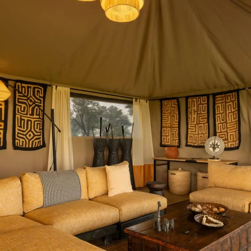 Interior of Ubuntu Migration Camp's lounge featuring cushioned sofas, woven textiles and wall art, soft pendant lighting, a small bar area, and open canvas walls overlooking surrounding woodland.