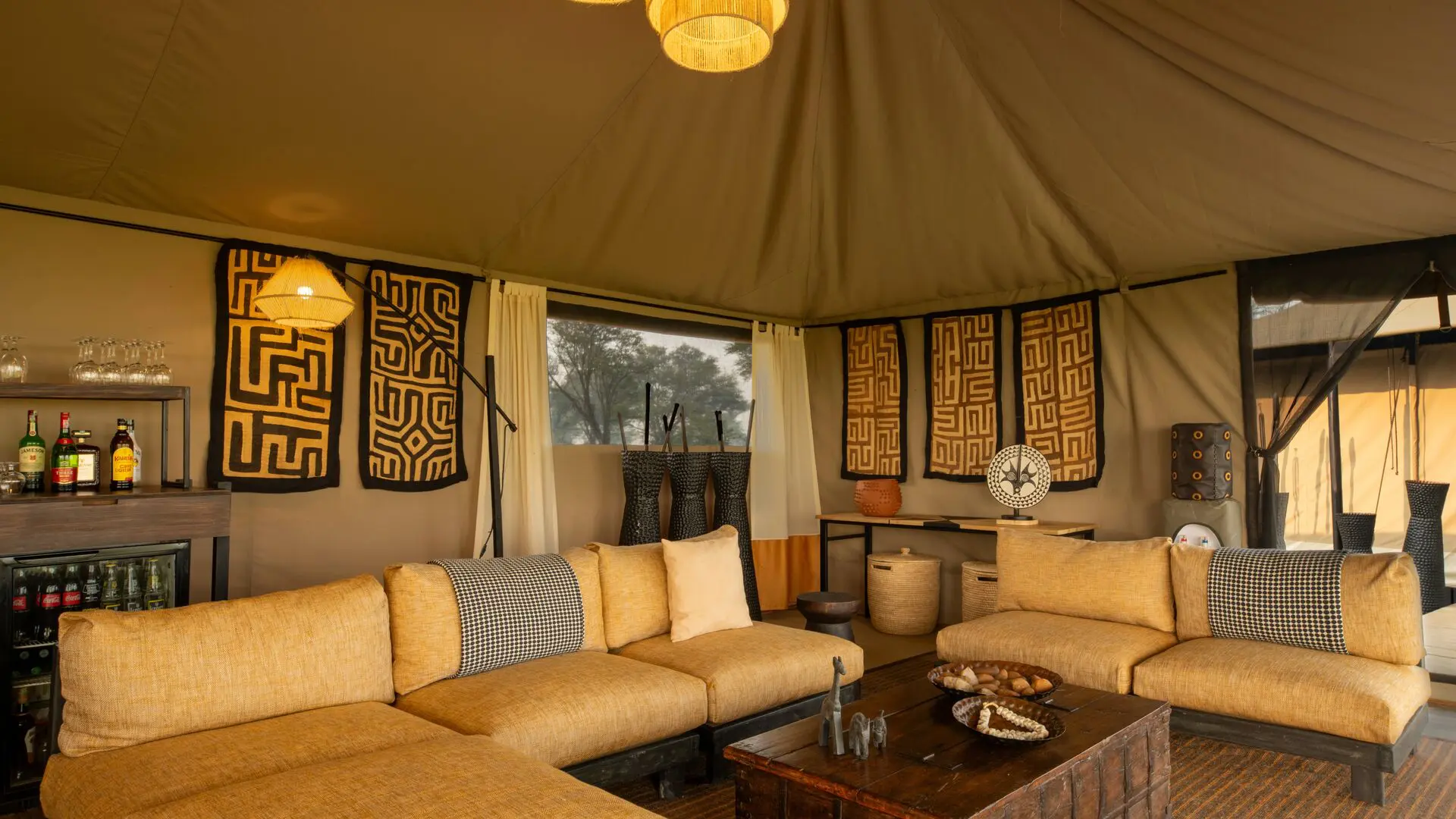 Interior of Ubuntu Migration Camp's lounge featuring cushioned sofas, woven textiles and wall art, soft pendant lighting, a small bar area, and open canvas walls overlooking surrounding woodland.