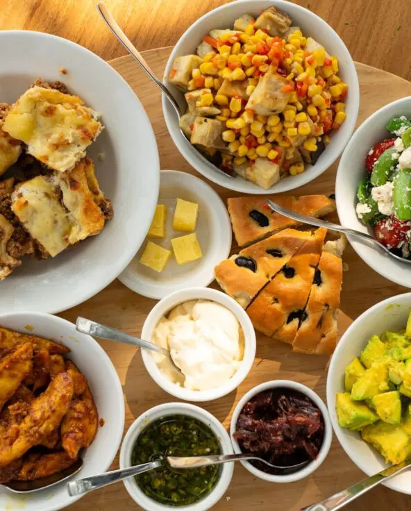 Overhead view of a safari lunch spread on a wooden table, featuring lasagne, roasted chicken, avocado salad, green bean and feta salad, corn and vegetable salad, focaccia bread, and assorted sauces in white bowls.