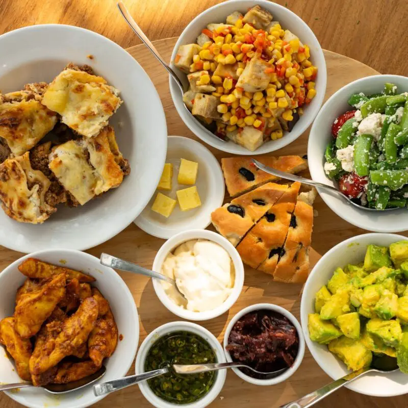 Overhead view of a safari lunch spread on a wooden table, featuring lasagne, roasted chicken, avocado salad, green bean and feta salad, corn and vegetable salad, focaccia bread, and assorted sauces in white bowls.