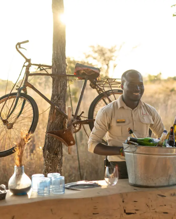 An Asilia staff member serving drinks at Usangu Camp's outdoor bar with bottles, glasses, and fruit on display