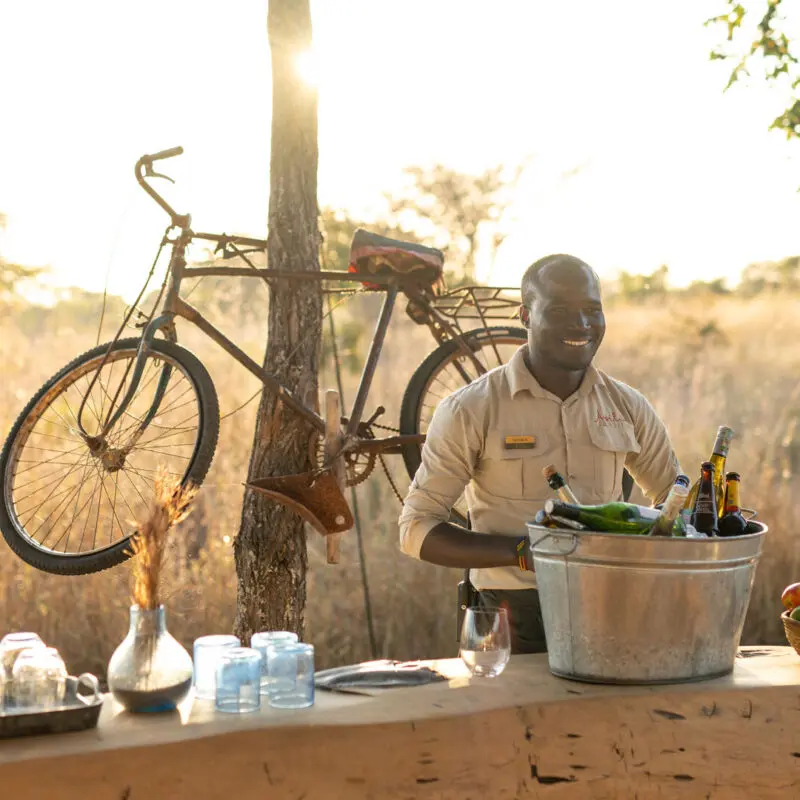 An Asilia staff member serving drinks at Usangu Camp's outdoor bar with bottles, glasses, and fruit on display