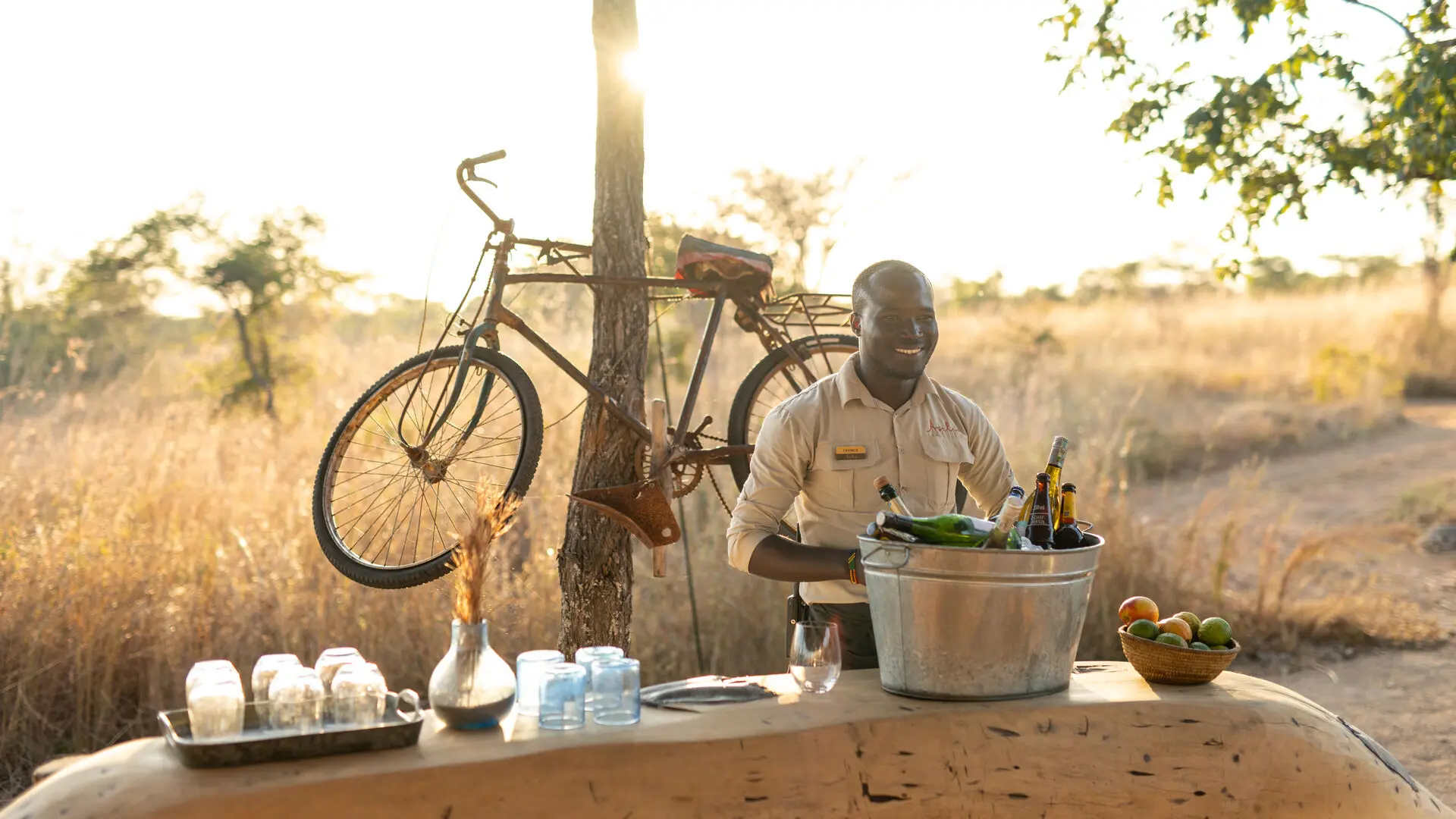 An Asilia staff member serving drinks at Usangu Camp's outdoor bar with bottles, glasses, and fruit on display