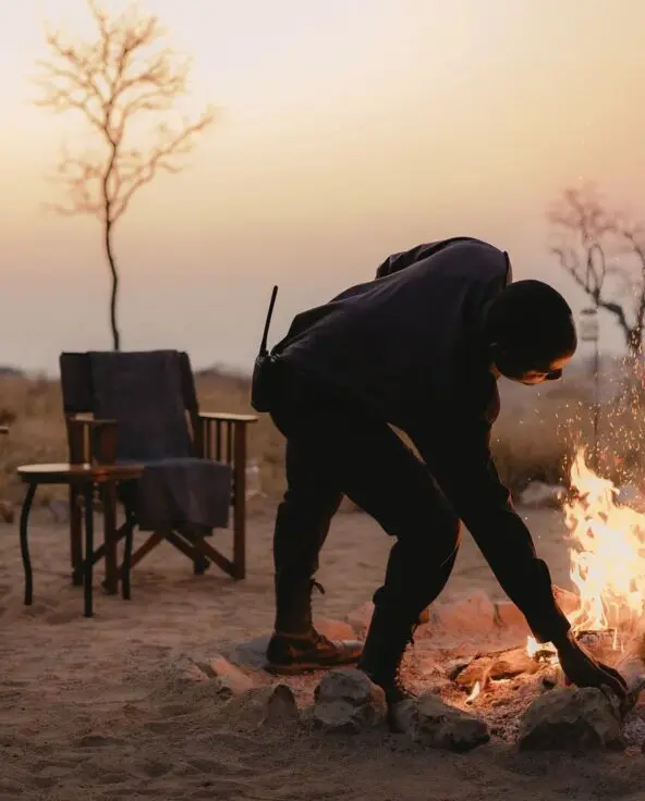 An Asilia staff member tends to a campfire with chairs set around in a dry landscape at dusk