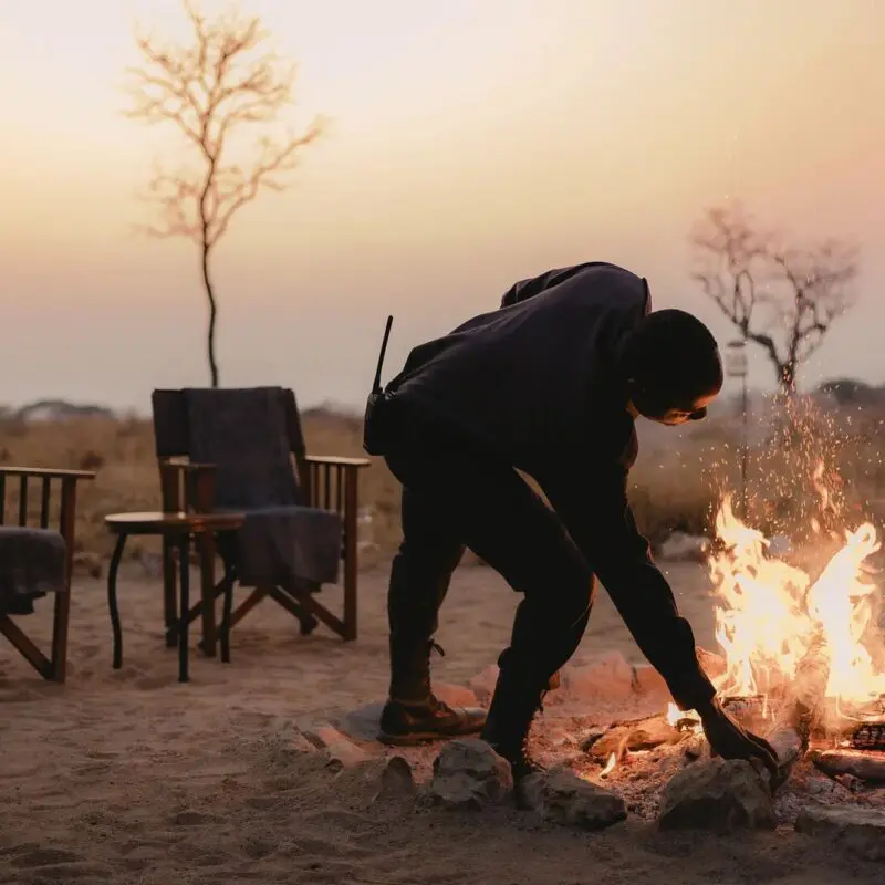 An Asilia staff member tends to a campfire with chairs set around in a dry landscape at dusk