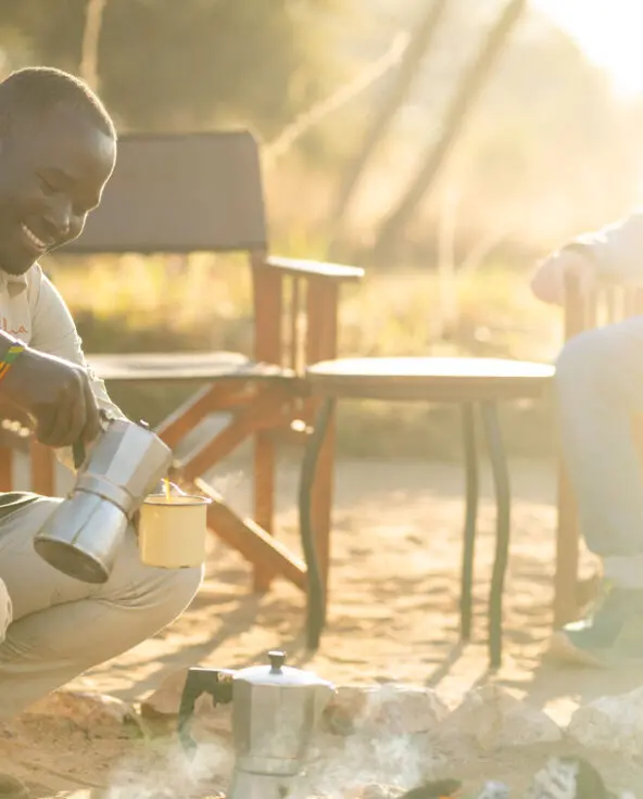 An Asilia staff member pouring coffee from a metal pot near a campfire with a guest seated nearby