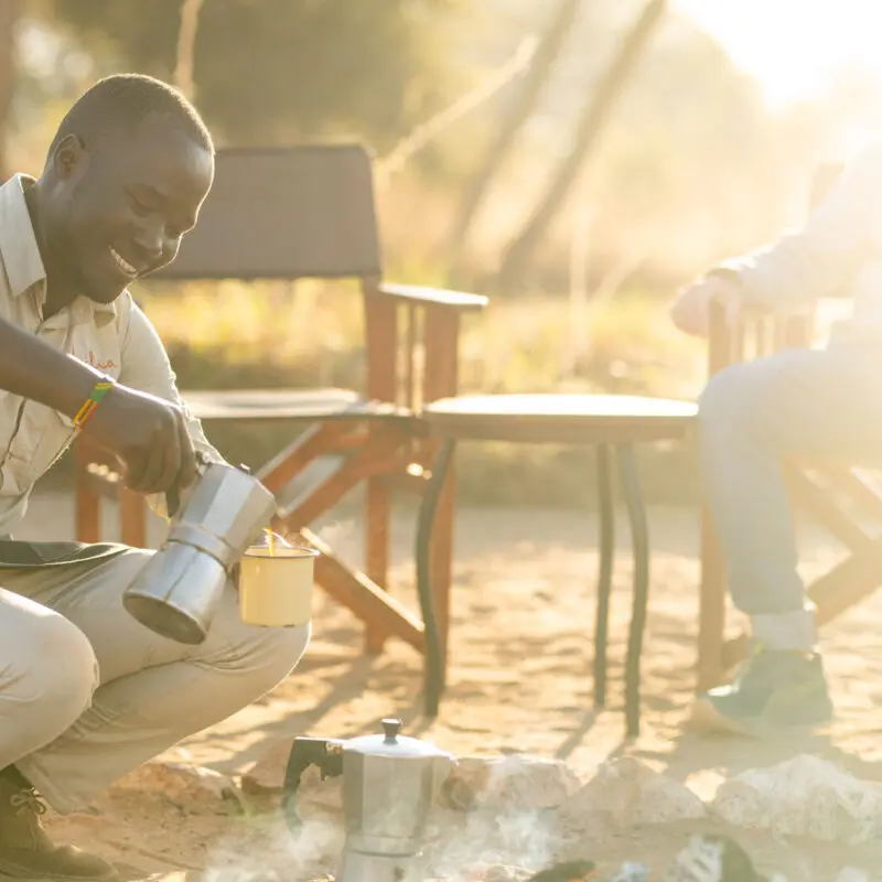 An Asilia staff member pouring coffee from a metal pot near a campfire with a guest seated nearby
