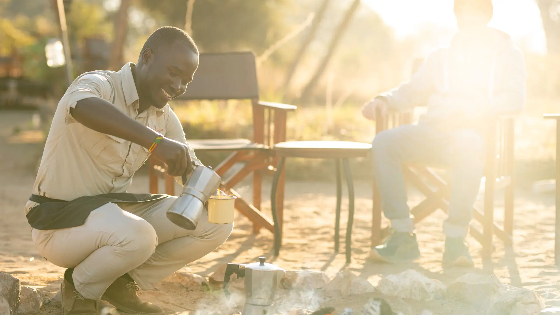 An Asilia staff member pouring coffee from a metal pot near a campfire with a guest seated nearby