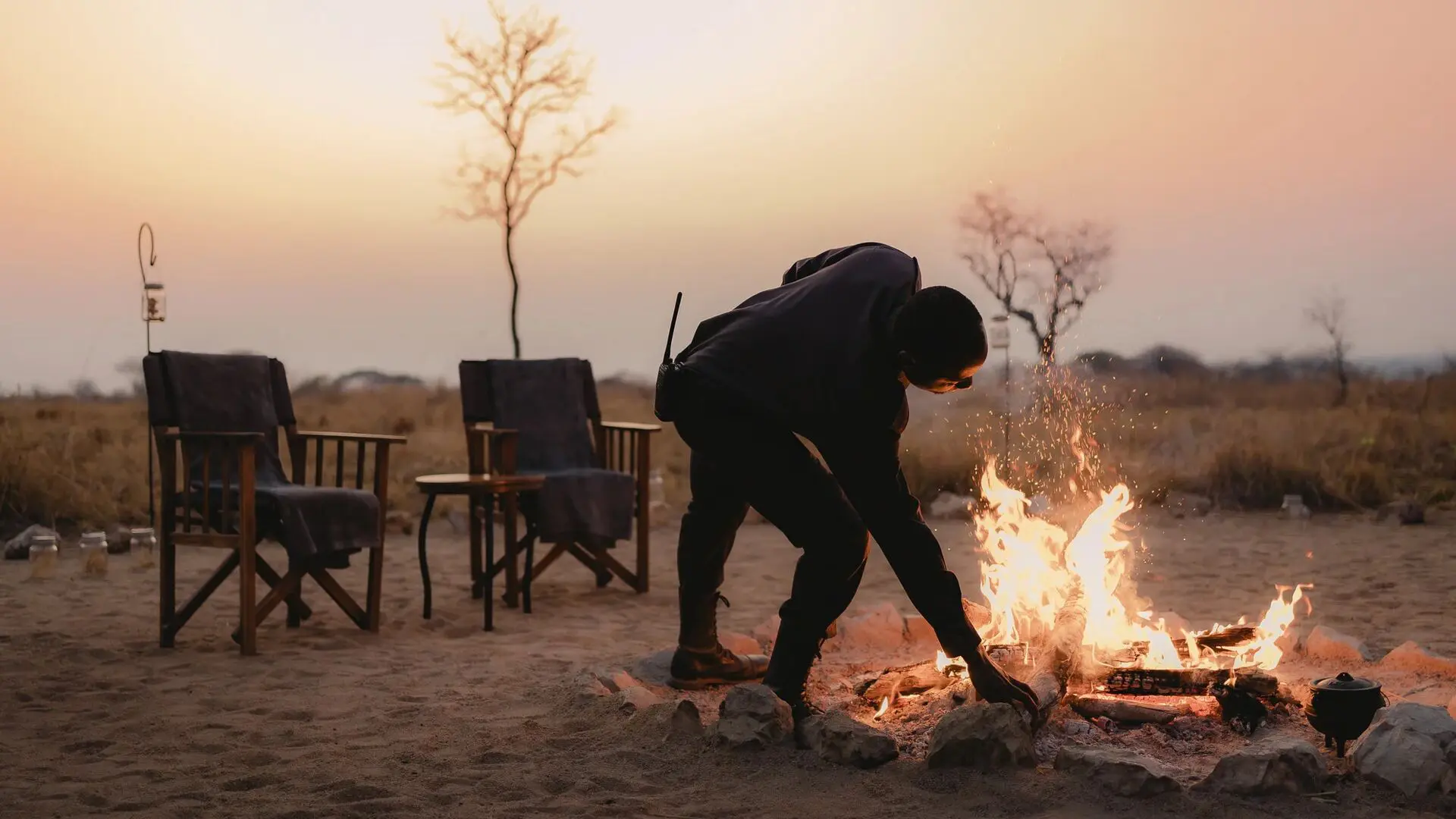 An Asilia staff member tends to a campfire with chairs set around in a dry landscape at dusk
