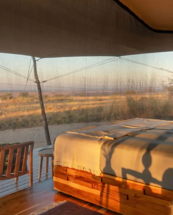 The interior of a guest bedroom at Usangu Camp with a bed facing expansive views of Ruaha National Park through mesh walls