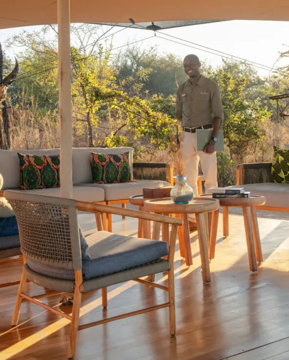 A guest seated in an open-air lounge speaking with a staff member among trees and soft morning light