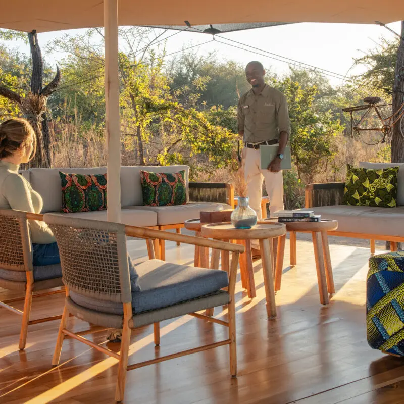 A guest seated in an open-air lounge speaking with a staff member among trees and soft morning light