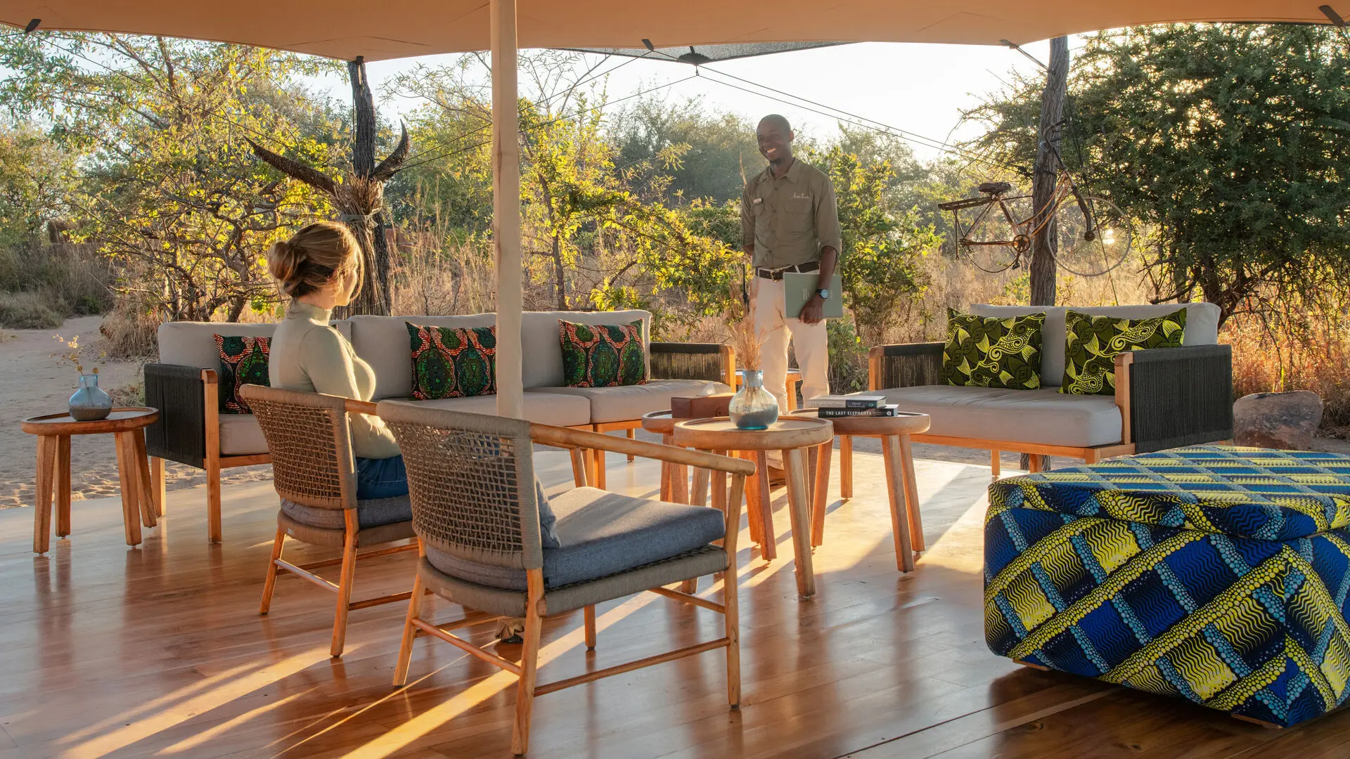 A guest seated in an open-air lounge speaking with a staff member among trees and soft morning light