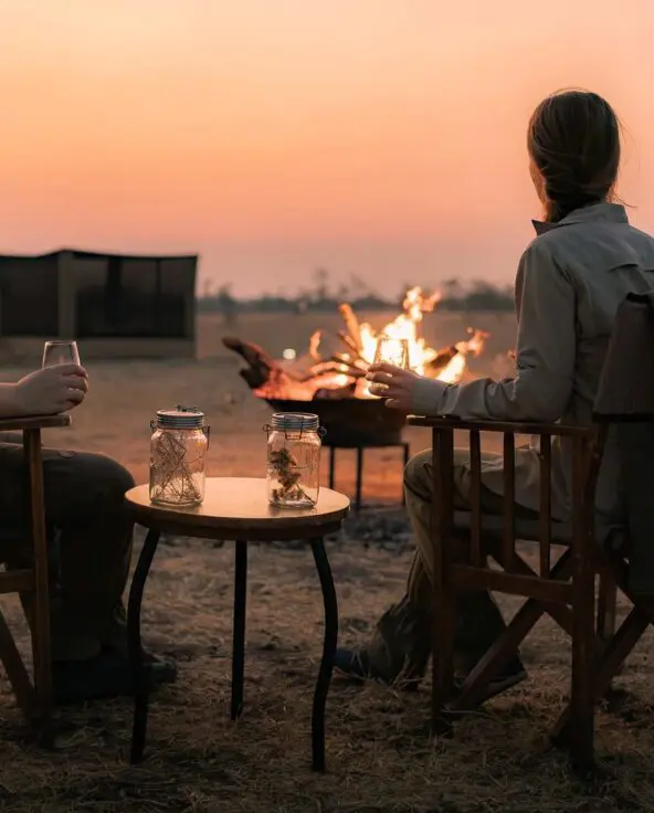 Two Asilia guests seated by a campfire at sunset, holding drinks and watching the fading light.