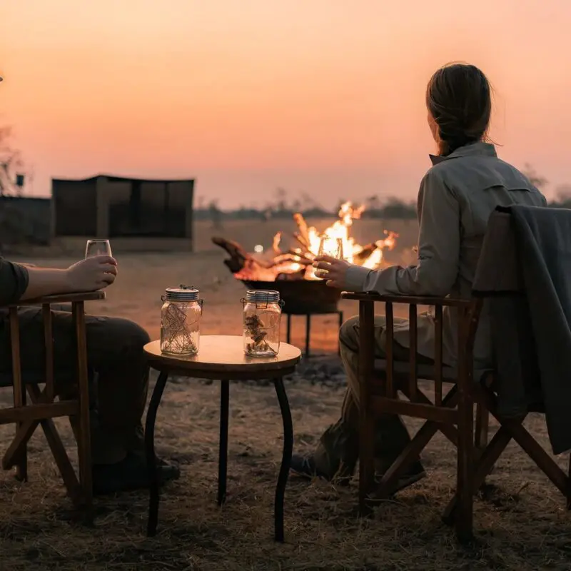 Two Asilia guests seated by a campfire at sunset, holding drinks and watching the fading light.