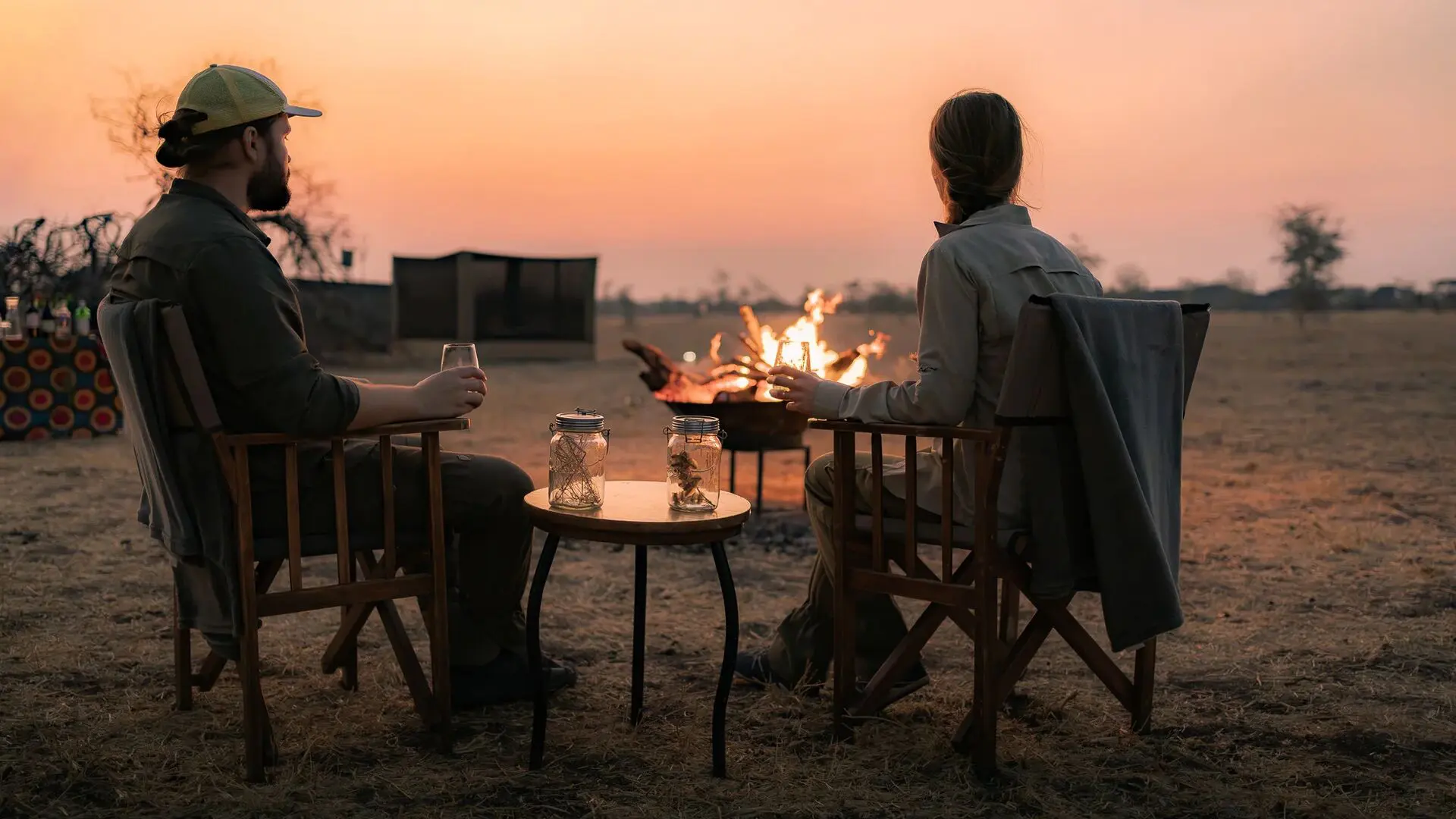 Two Asilia guests seated by a campfire at sunset, holding drinks and watching the fading light.