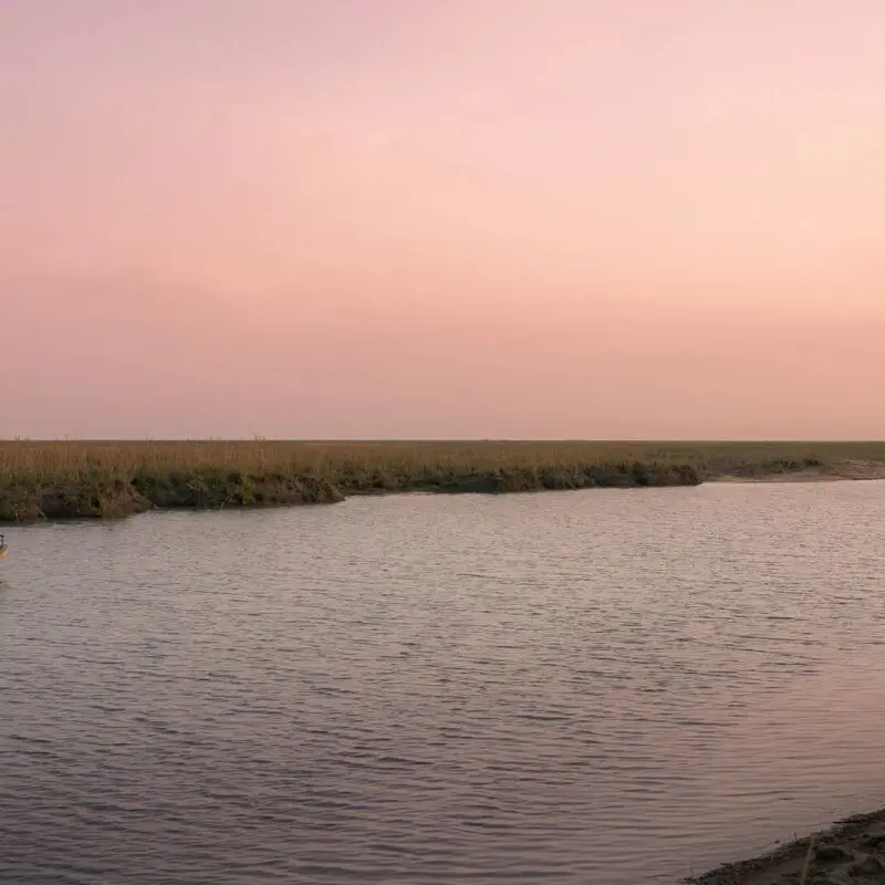 A small boat with safari guests traveling along a calm river in Ruaha National Park under a soft pink sky