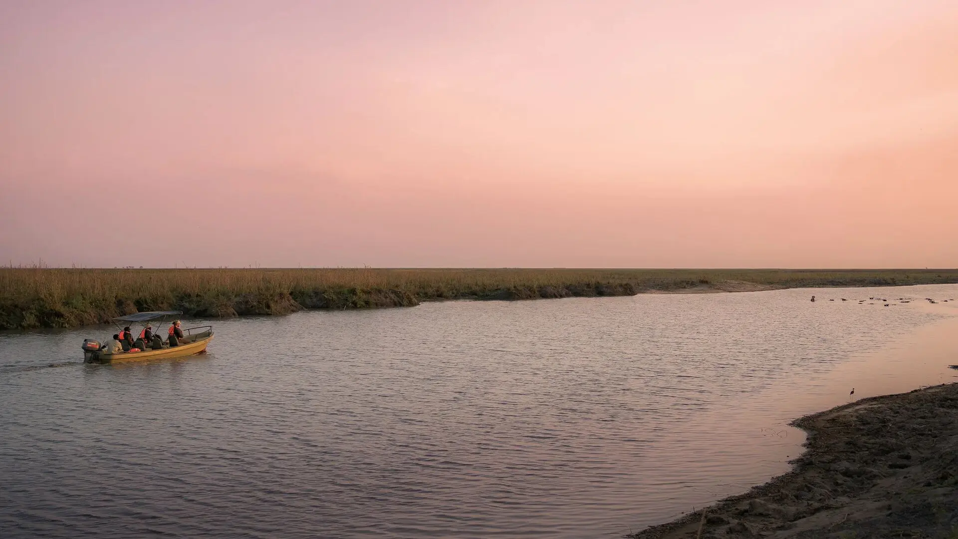 A small boat with safari guests traveling along a calm river in Ruaha National Park under a soft pink sky
