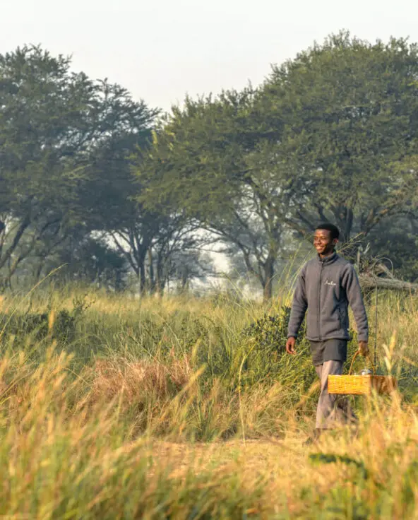 Smiling Asilia staff member walks through long grass carrying a basket