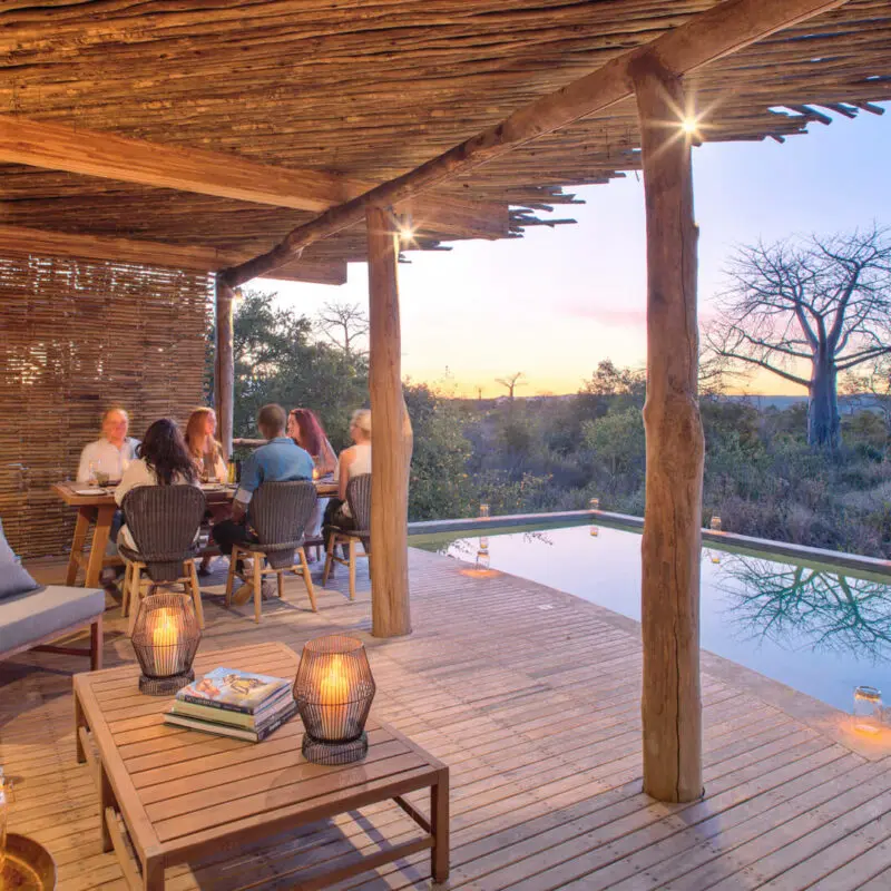 Jabali Private House, Ruaha National Park, guests having dinner by the pool lit up with lanterns at sunset, deck loungers, table with books, and candles