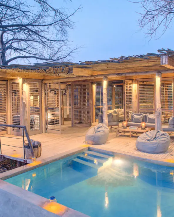 Jabali Private House, Ruaha National Park, house lit up with lanterns once the sun has set, blue pool, tables, chairs, loungers and bean bags for seating, shutter doors open