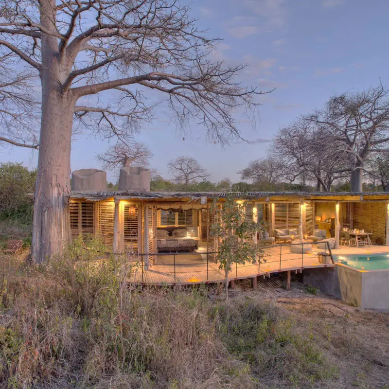 Jabali Private House, Ruaha National Park, view of the outside at sunset with pool and lanterns lighting up the deck with tables and chairs and open shutters to the room