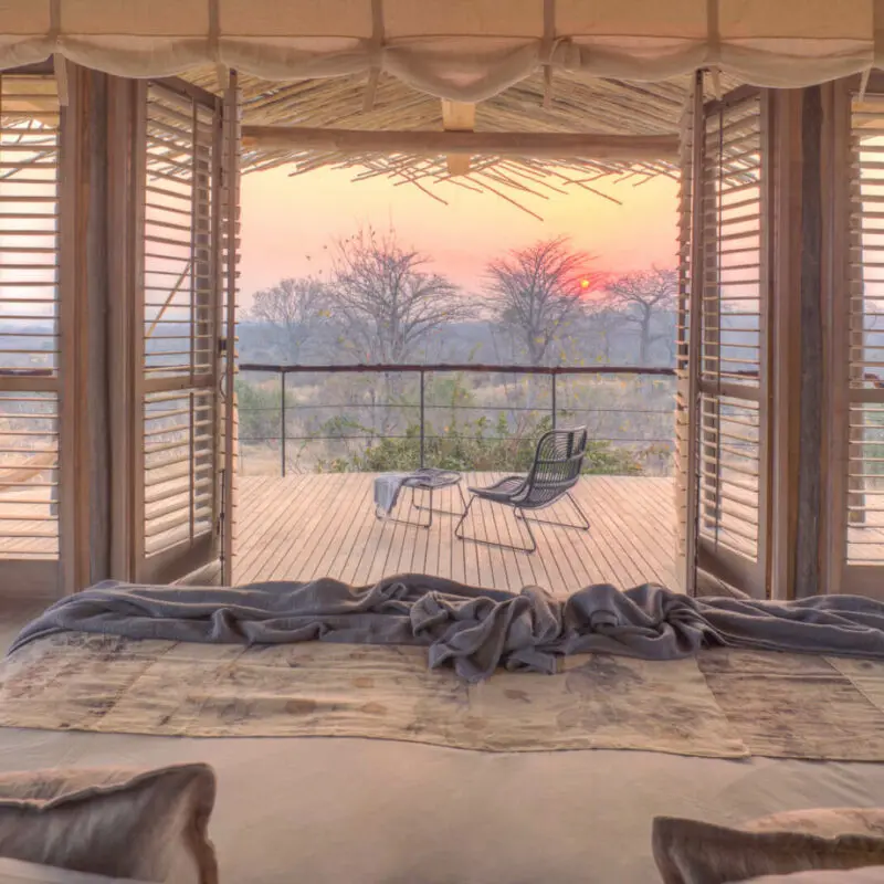 Jabali Ridge, Ruaha National Park, double bed with pillows and throw blanket, open shutters leading out to the deck with table and chair