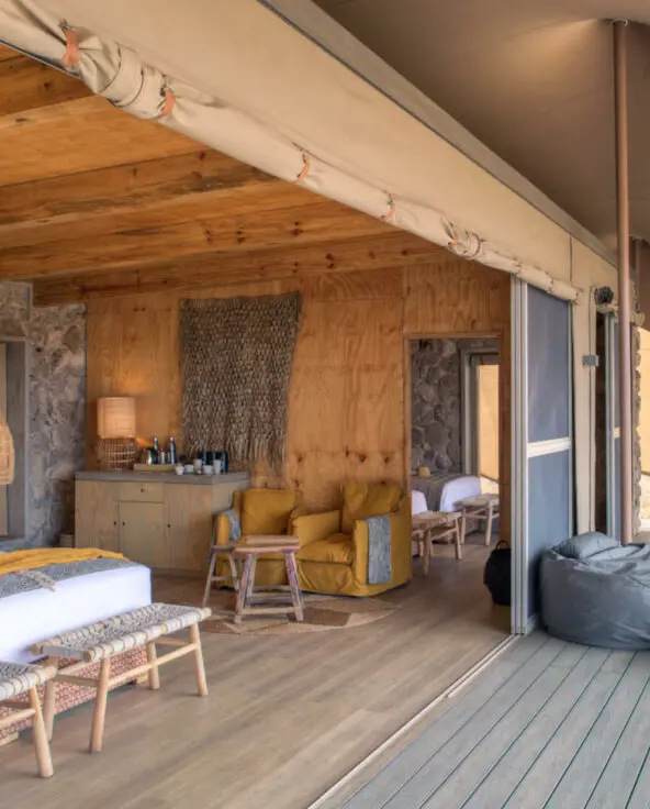 Namiri Plains, Serengeti National Park, view of the interior, double guest tent, bed with pillows and throw blanket, hanging lights, lamp on the side table with coffee station, bean bags on the deck, doors open