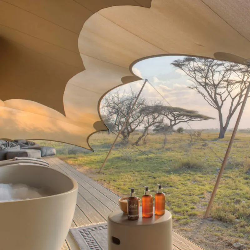 Namiri Plains, Serengeti National Park, Bath on the deck with bubbles and wine glass and bottle, amenity bottles and bath salts, loungers and bean bags