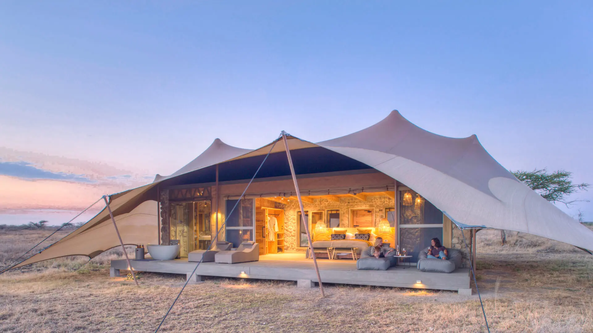 Namiri Plains, Serengeti National Park, guests having wine on the deck outside the tent, open doors, lights on and loungers on the deck with bath