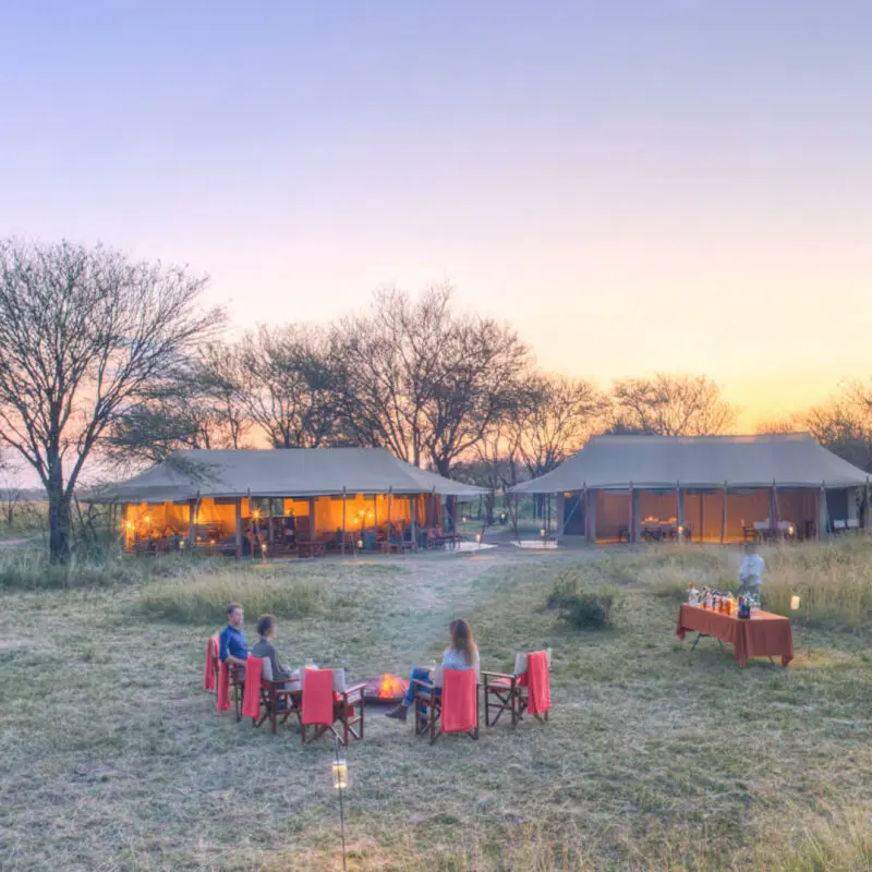 Guests enjoying sundowners around the campfire at camp, olakira migration camp, Serengeti National Park, Tanzania