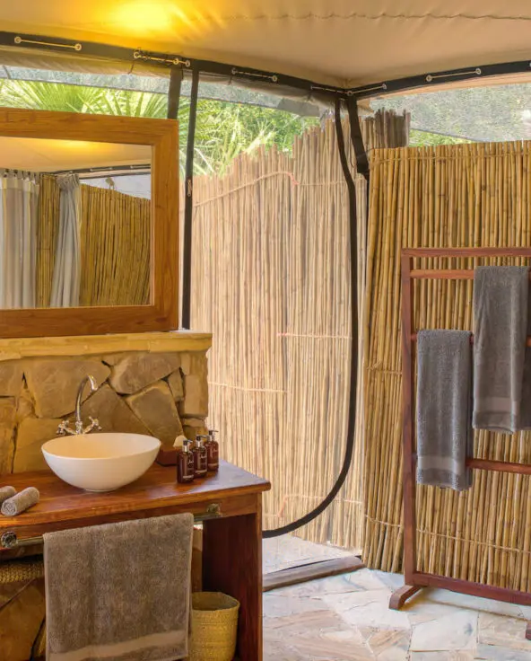 Roho ya Selous Camp, Nyerere National Park, guest bathroom with double vanity and towels hanging on a rack near a plant, bamboo surrounding