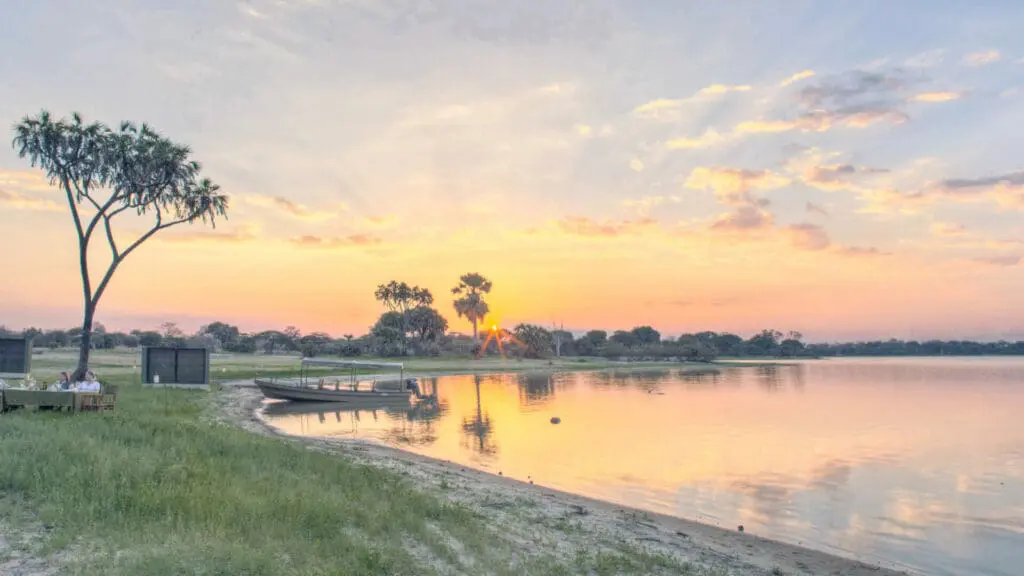Roho ya Selous Camp, Nyerere National Park, guests seated on the banks of the river with a boat on the shore, sun setting in the back
