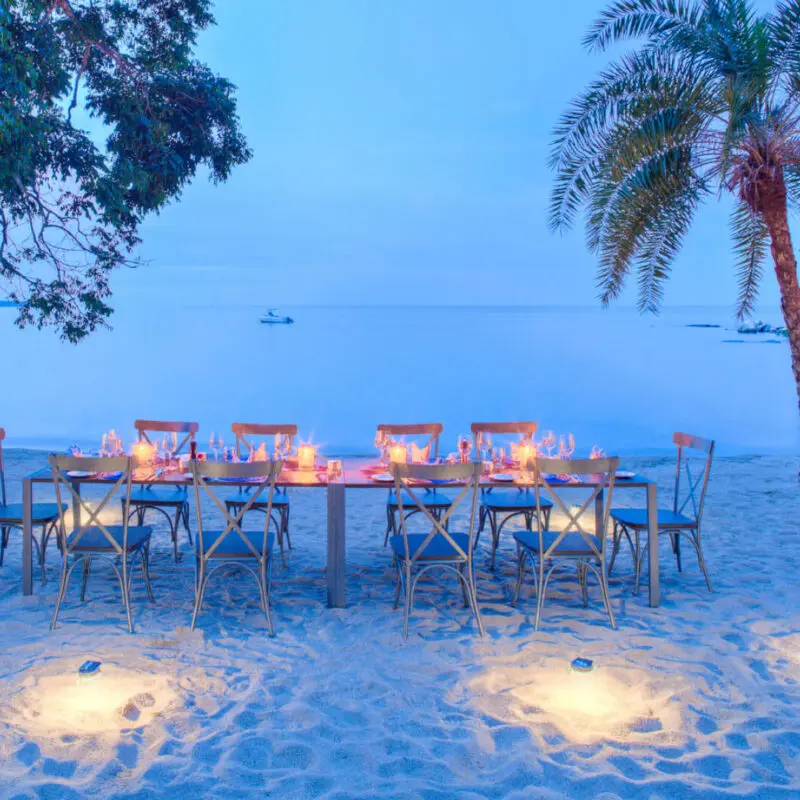Rubondo Island Camp, Lake Victoria, private dining on the shore, setup with candles at sunset, sand, palm trees and lake in the background