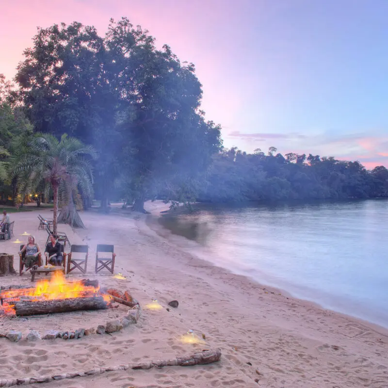 Rubondo Island Camp, Lake Victoria, guests enjoying sundowners around the firepit along the banks of Lake Victoria