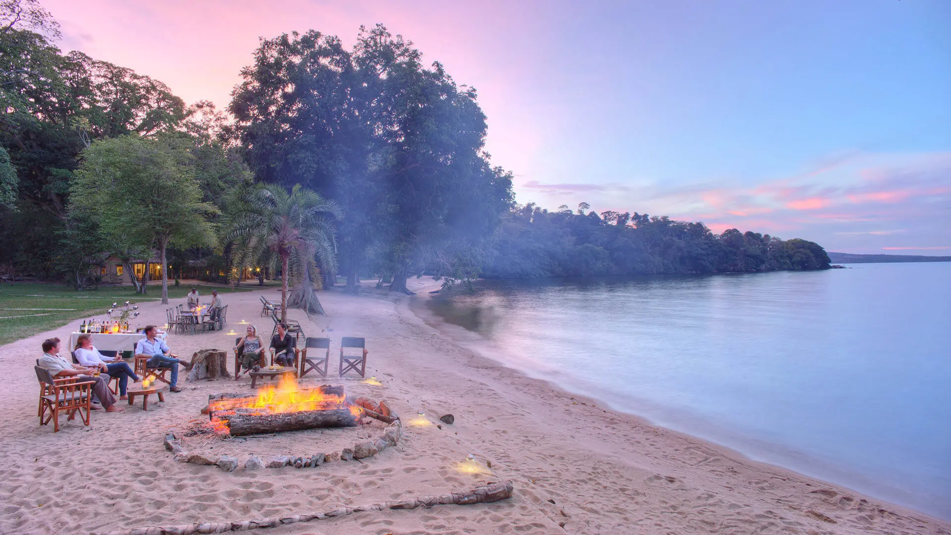 Rubondo Island Camp, Lake Victoria, guests enjoying sundowners around the firepit along the banks of Lake Victoria