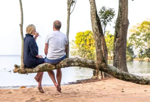 A couple enjoy a sundowner whilst sat on a branch on the beach at Rubondo Island Camp in Tanzania - Asilia Africa
