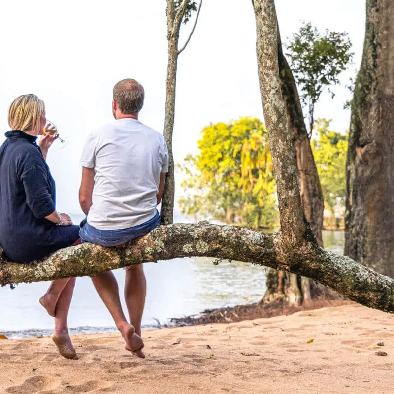 A couple enjoy a sundowner whilst sat on a branch on the beach at Rubondo Island Camp in Tanzania - Asilia Africa