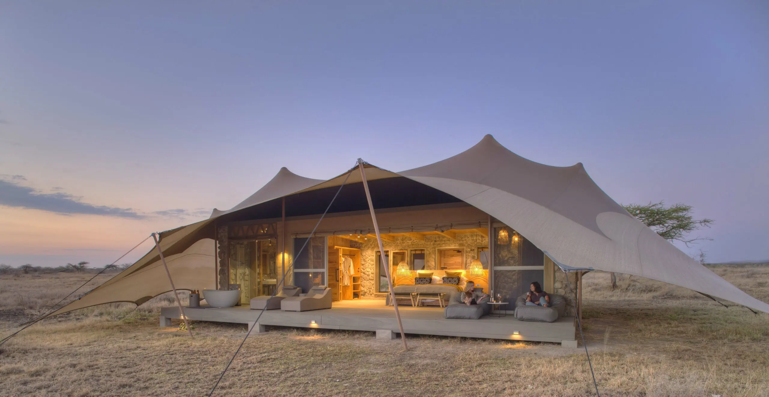Asilia Namiri Plains guest tent exterior, guest on the deck loungers enjoying wine, lights on inside, doors open