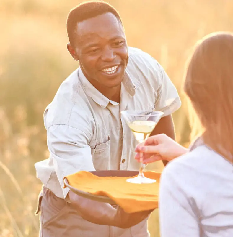 Smiling staff member serving a guest a drink at Olakira Migration Camp at sunset