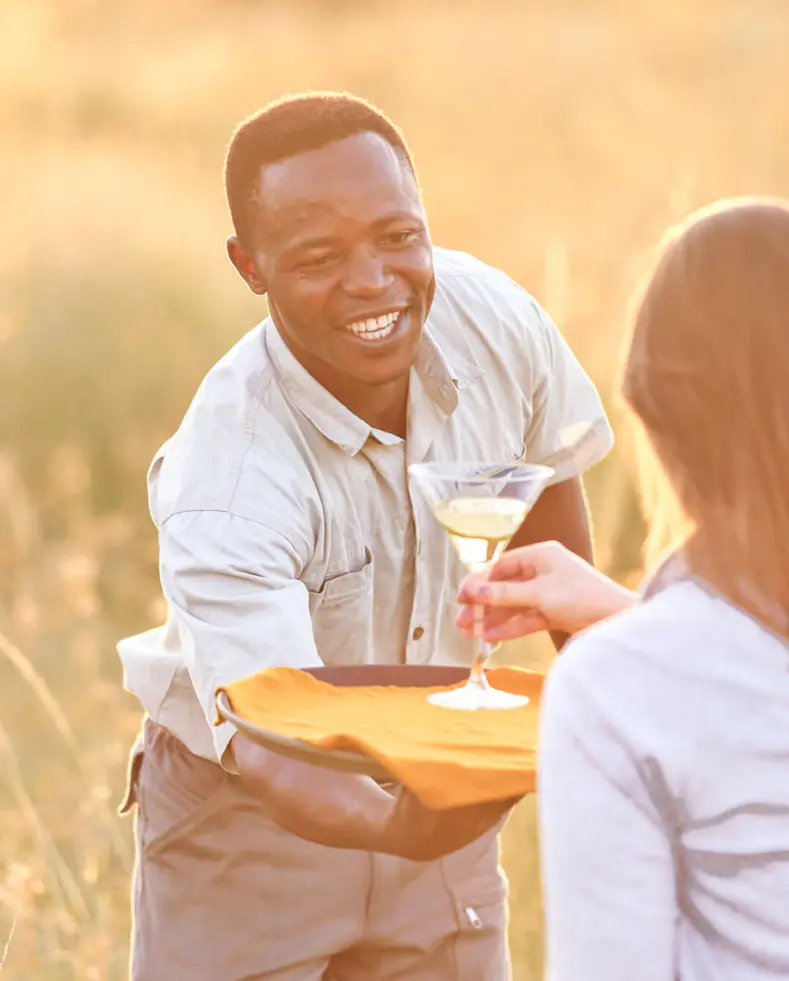 Smiling staff member serving a guest a drink at Olakira Migration Camp at sunset