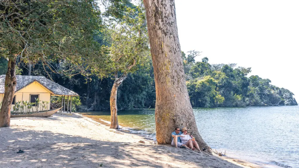 couple sitting under a tree on the waters edge, rubondo island, lake victoria, tanzania, asilia africa