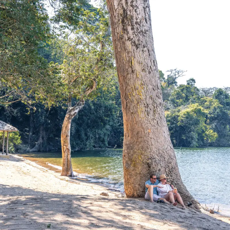 couple sitting under a tree on the waters edge, rubondo island, lake victoria, tanzania, asilia africa