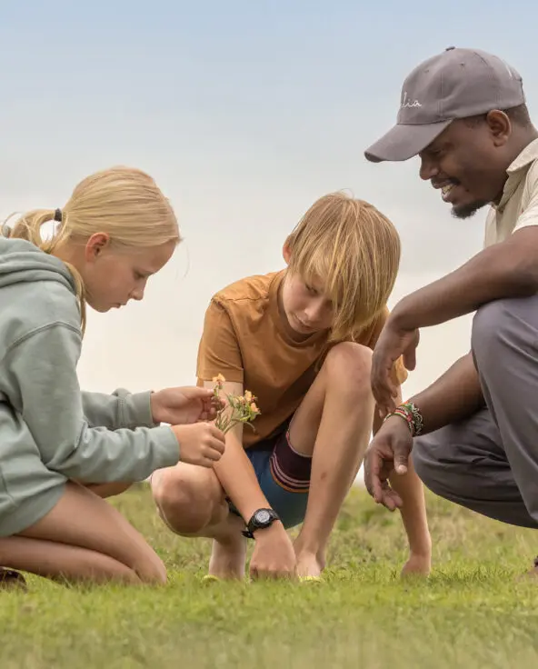 a male safari guide crouches with two children exploring nature at ol pejeta camp in kenya, asilia africa