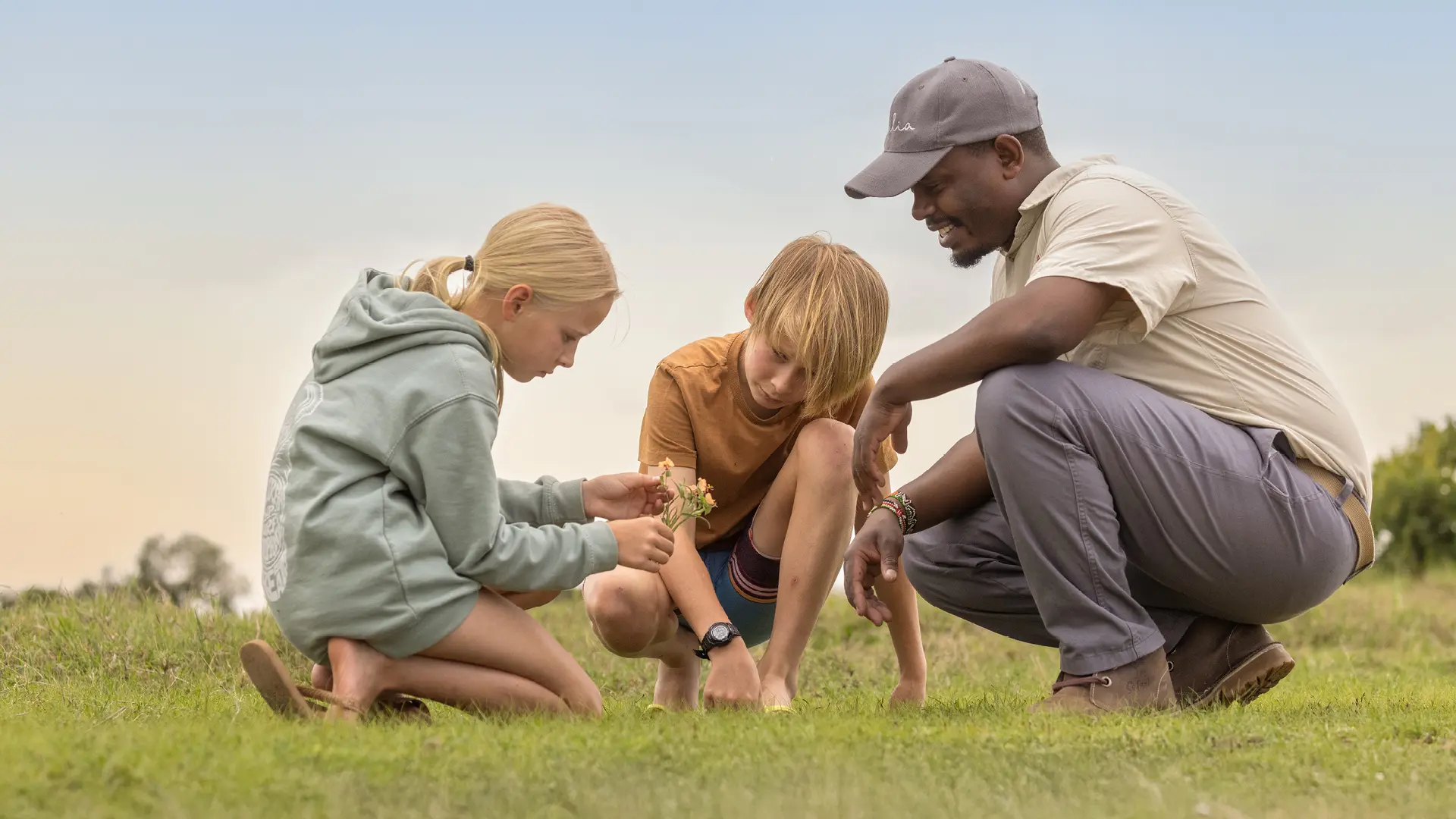 a male safari guide crouches with two children exploring nature at ol pejeta camp in kenya, asilia africa